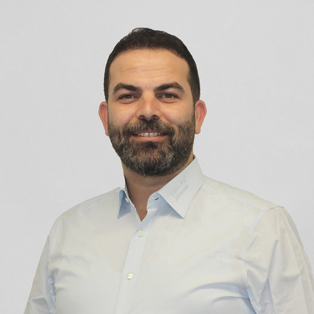 New Head of Sales SIMALFA® 1 Portrait of a smiling man with a dark beard and short hair, wearing a light blue shirt. The shirt collar subtly displays the embroidered text "SIMALFA Adhesives."