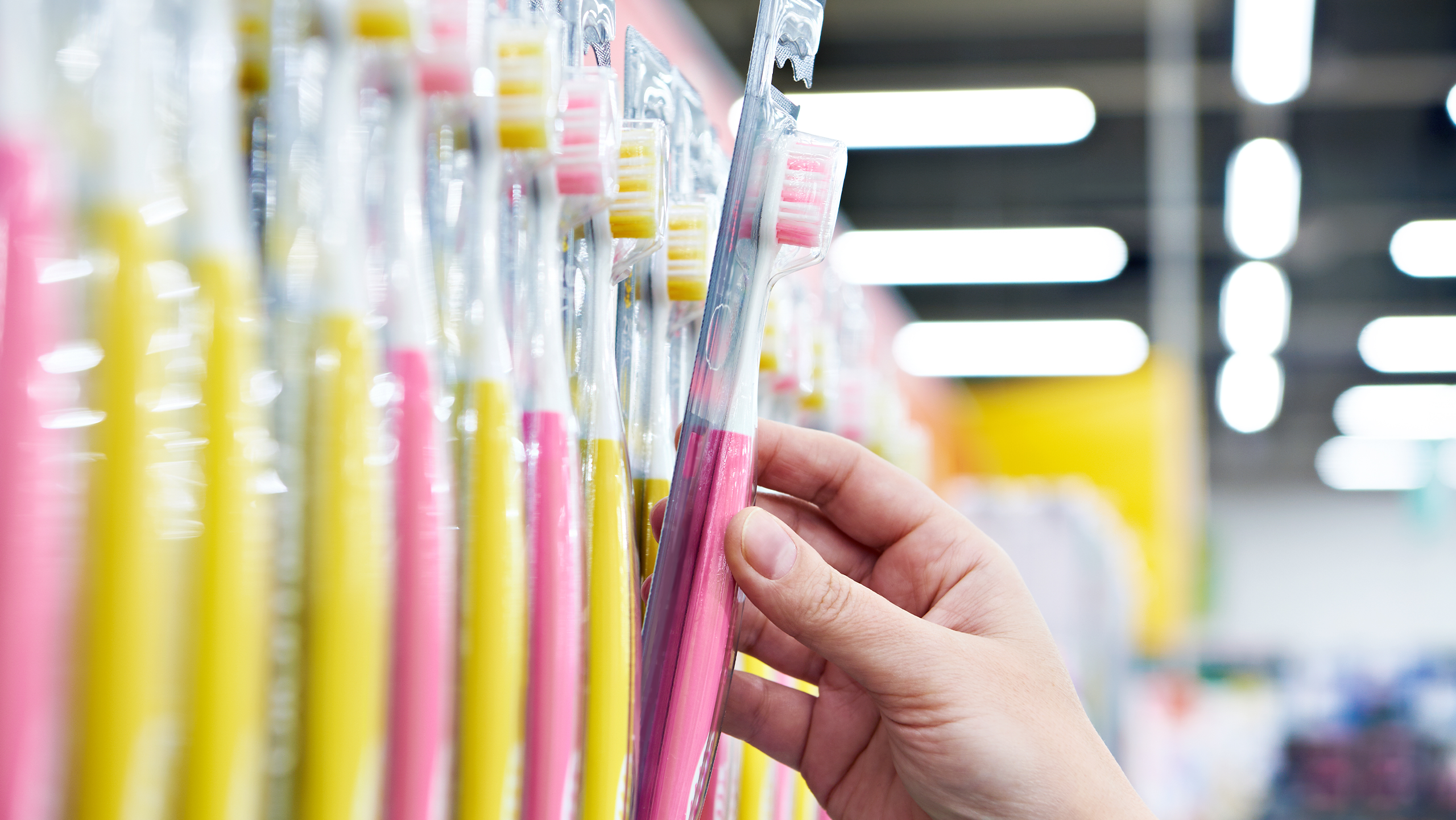 Hand greift nach einer Zahnbürste in einem Supermarktregal, umgeben von zahlreichen weiteren Zahnbürsten in Verpackungen.